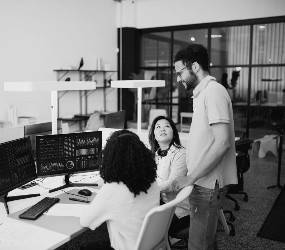 Three colleagues at a workstation reviewing data dashboards on multiple monitors.